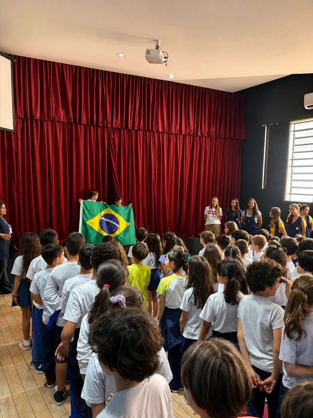 Alunos reunidos no palco em atividade de educação cívica com a bandeira do Brasil